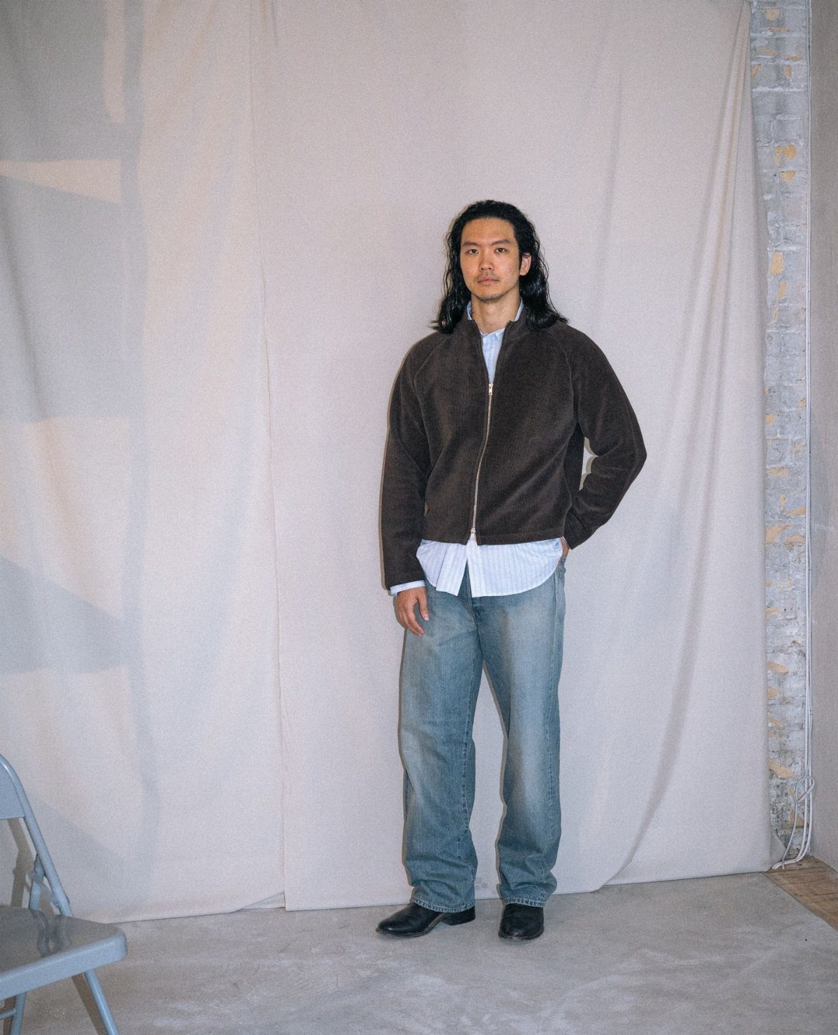 A man posing in a brown jacket, striped shirt, and baggy blue jeans.