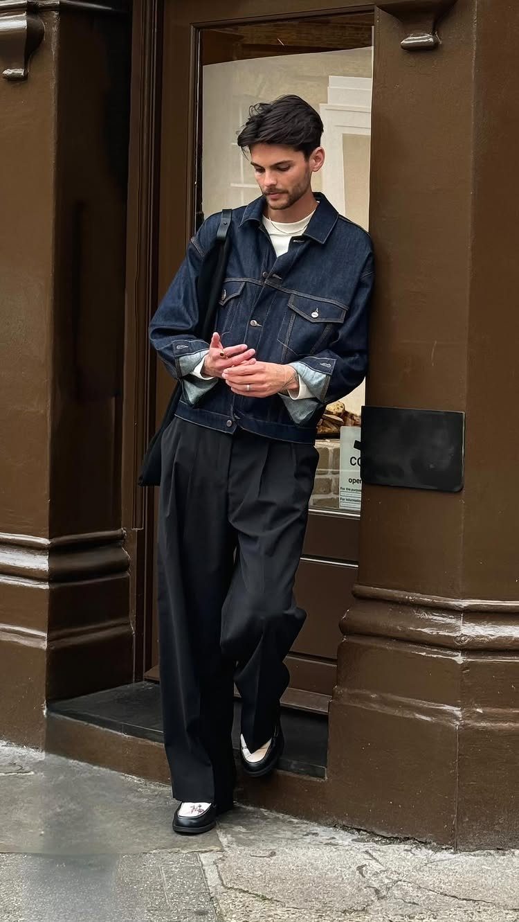 Man in a denim jacket and wide-leg black trousers leaning against a dark brown doorway.