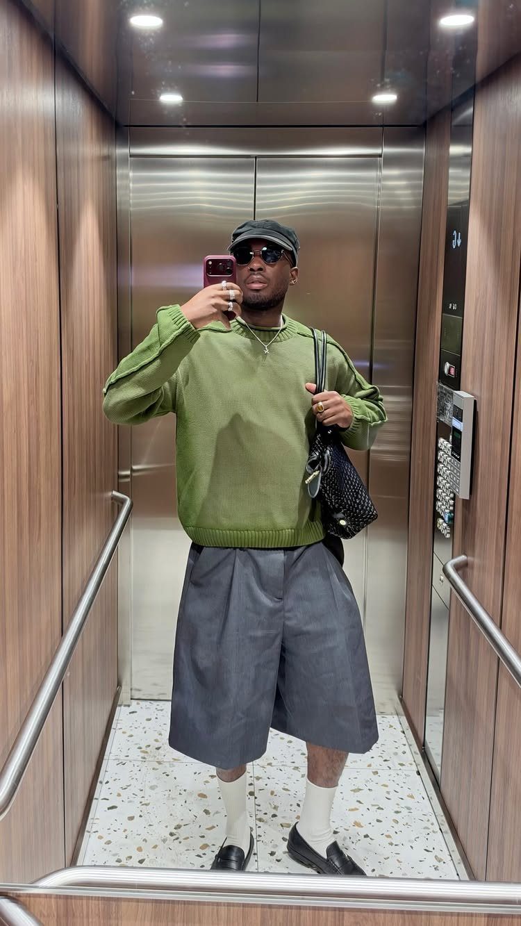 Man in an elevator wearing an olive green sweater, grey shorts, and black loafers.