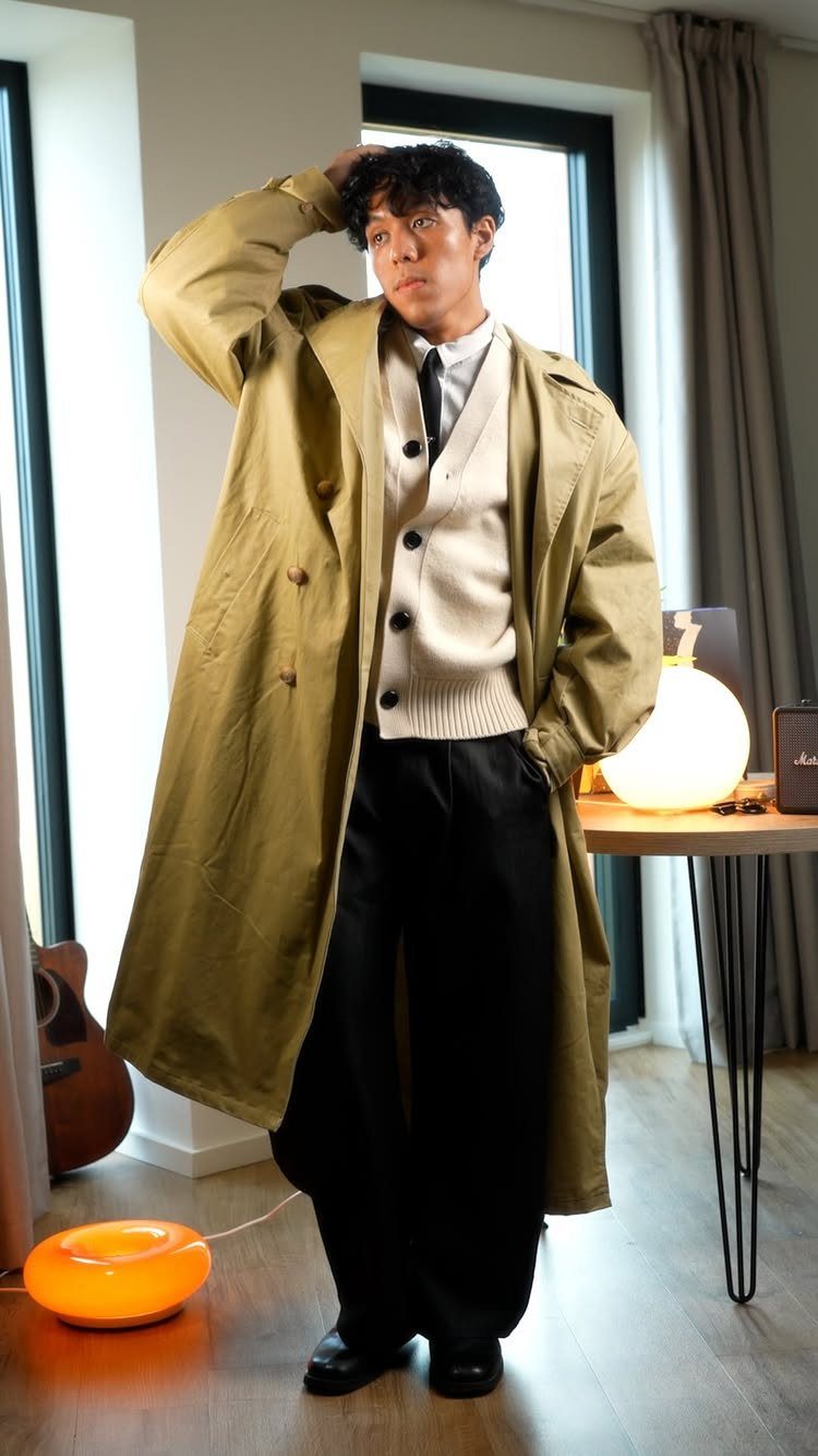 Man wearing a tan trench coat layered over a cream cardigan and black trousers.