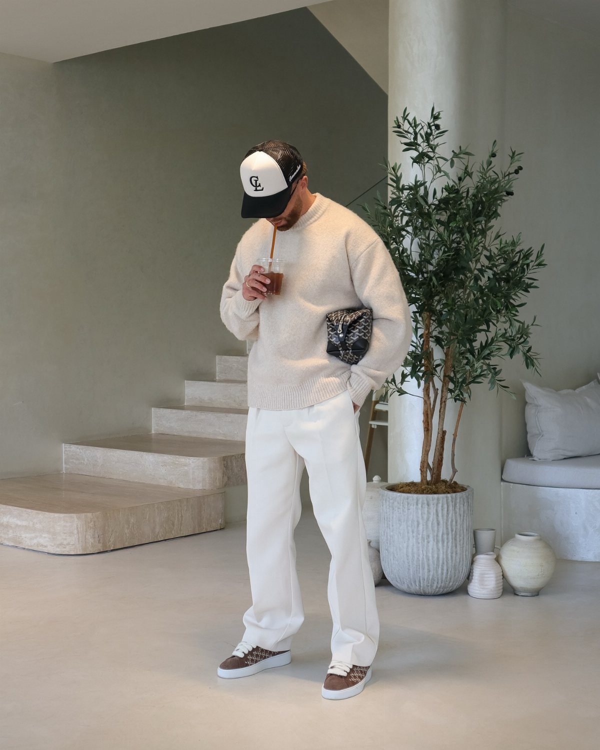 Man in cream sweater and white trousers standing in a minimalist neutral-toned room.