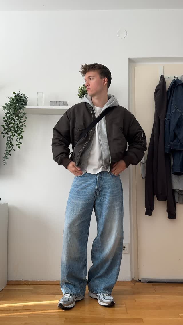 Young man posing in a dark bomber jacket, grey hoodie, and oversized wide-leg blue jeans.