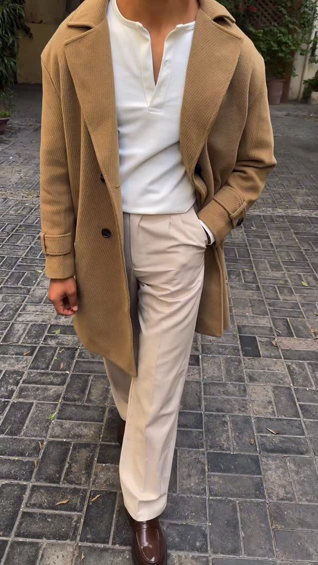 Man wearing a tan overcoat, white henley, and beige trousers standing on cobblestones.