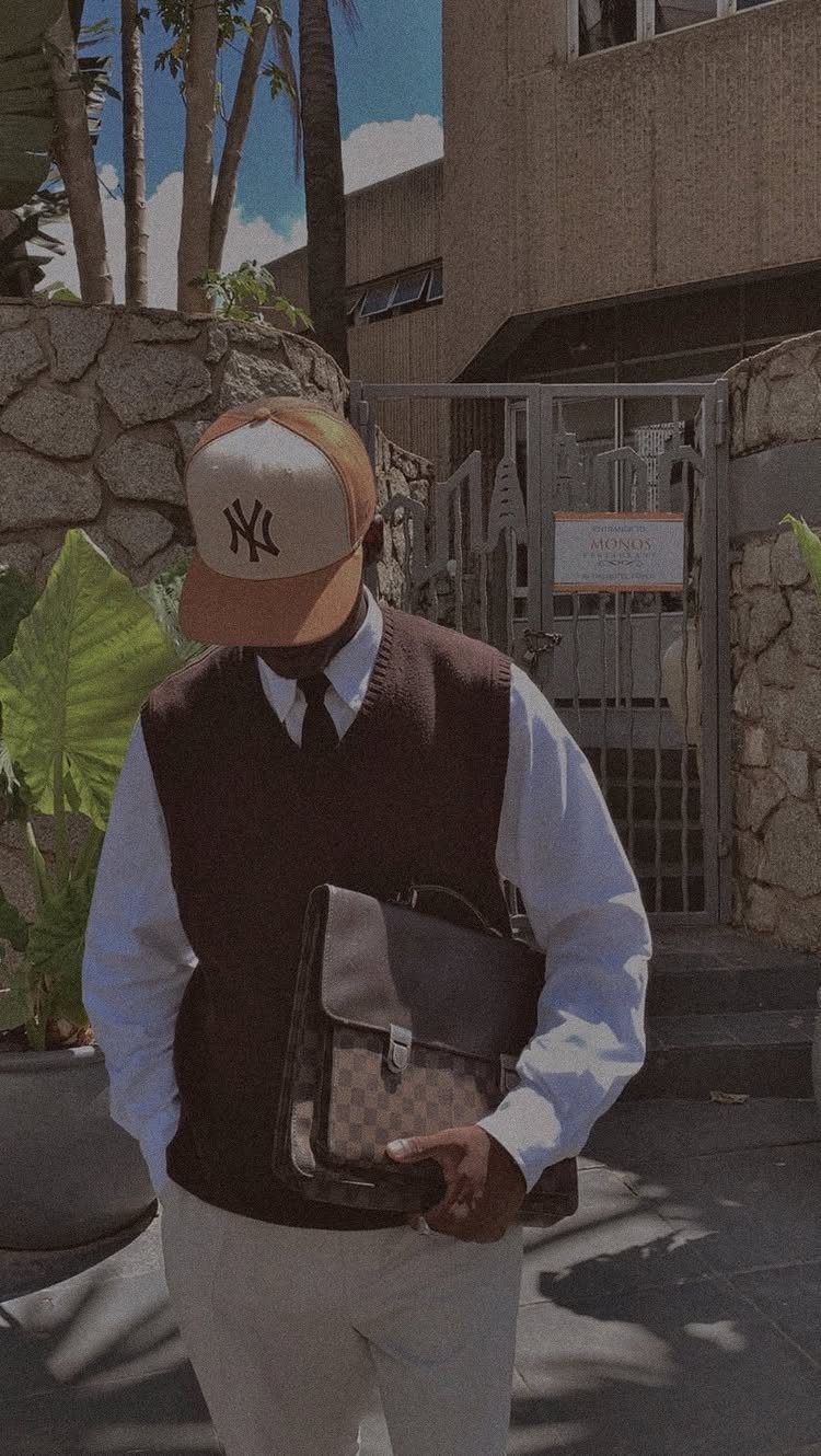 A man wearing a sweater vest, tie, baseball cap, and holding a briefcase outdoors.