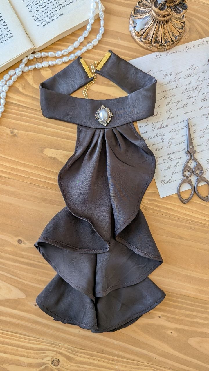 A ruffled dark grey silk cravat with a pearl brooch resting on a wooden table.