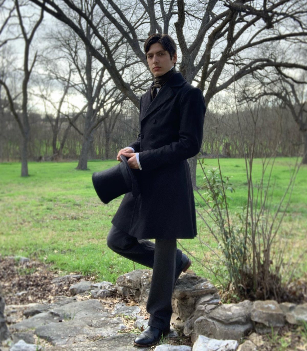 Man in a black Victorian frock coat and trousers standing on a stone wall outdoors.