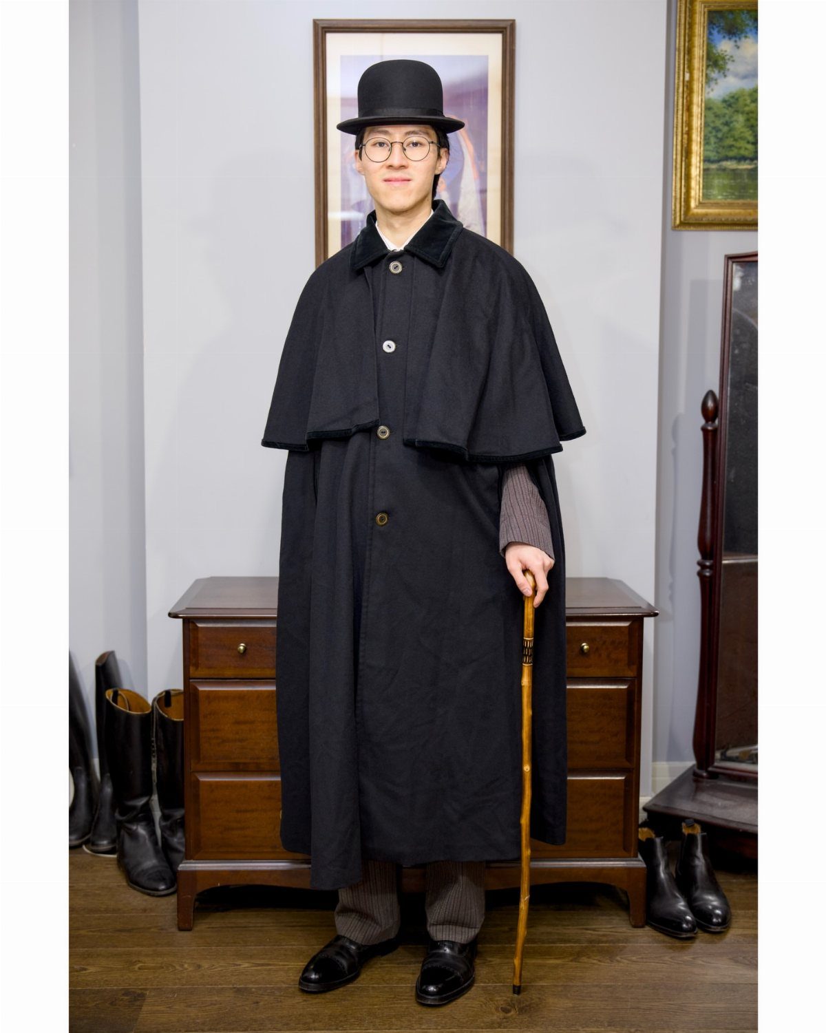 A man in a black bowler hat and long black Inverness cape holding a wooden cane.