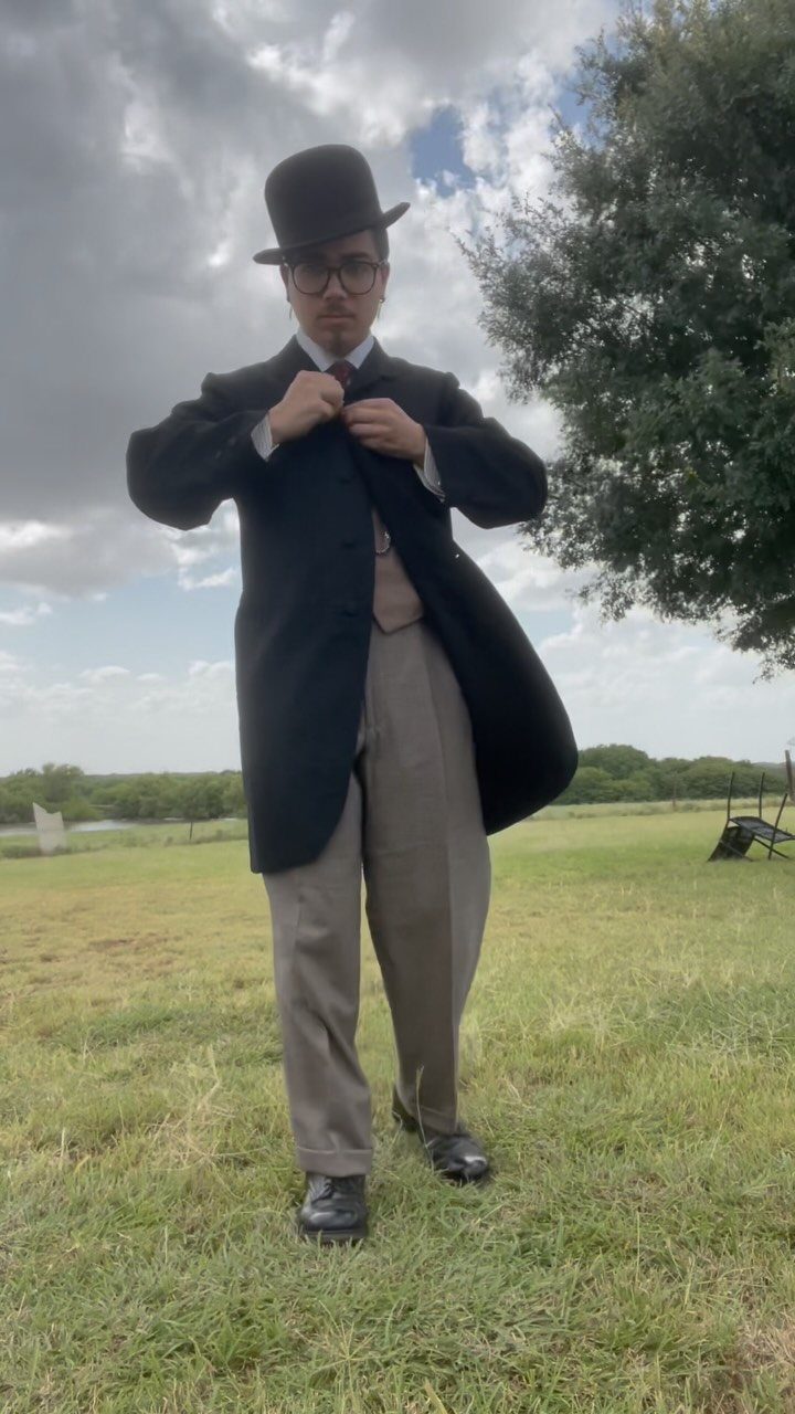 A man in a black morning coat and top hat standing in a grassy field.