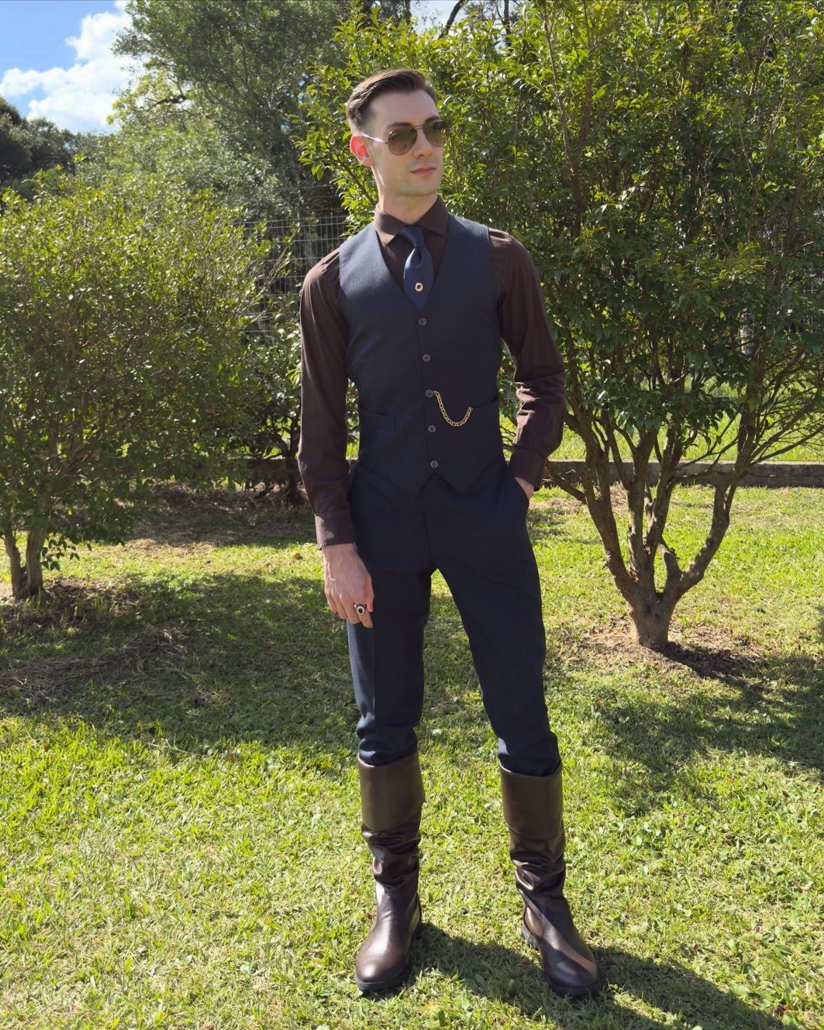 Man in Victorian-style waistcoat and trousers wearing tall brown leather boots standing on grass.