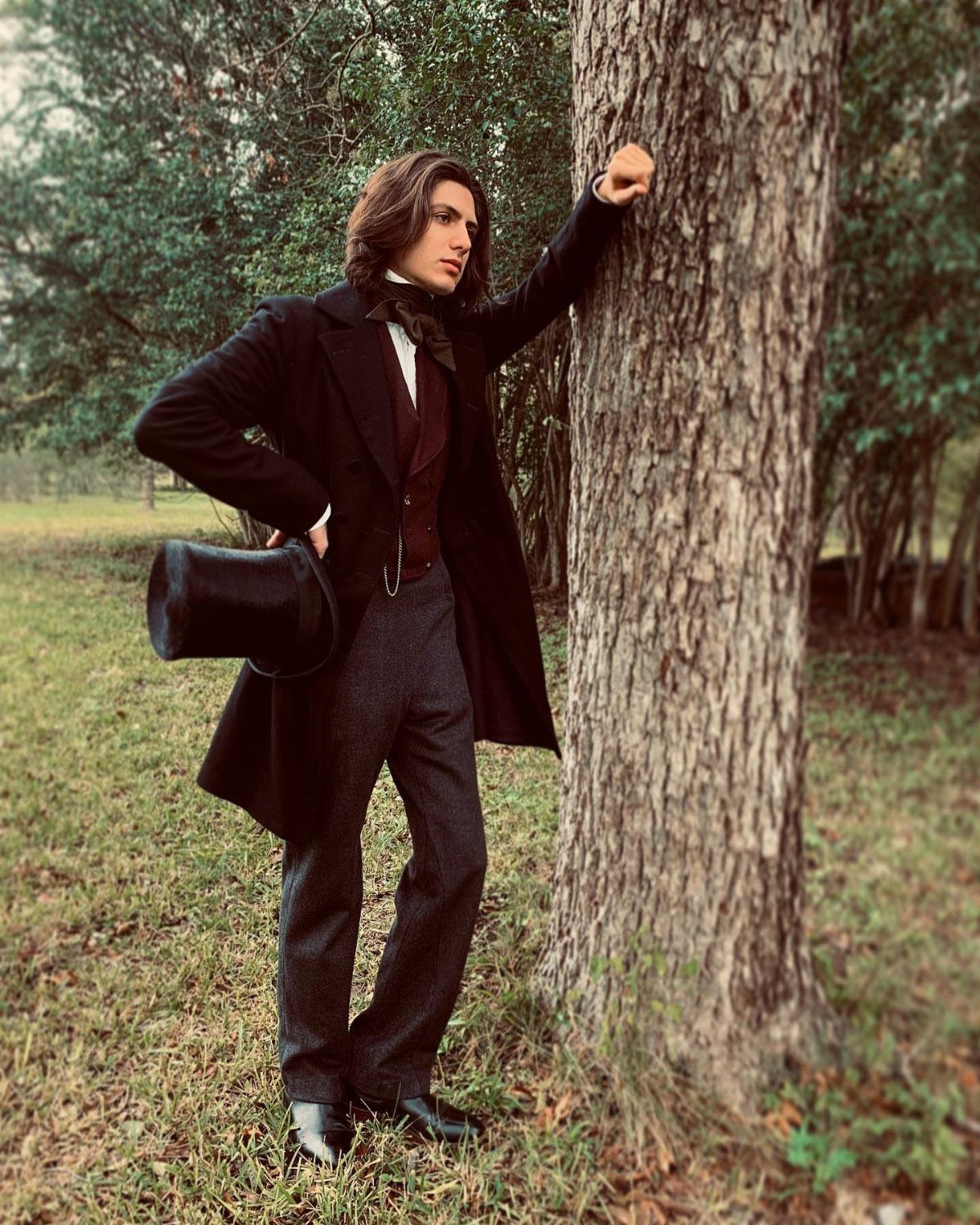 A man in a dark Victorian frock coat and waistcoat leaning against a tree.