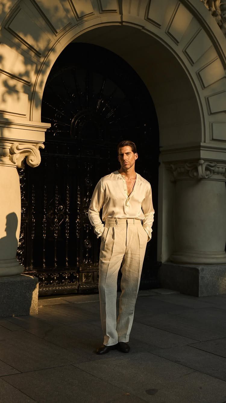Man in a cream silk shirt and matching trousers standing before a stone archway.