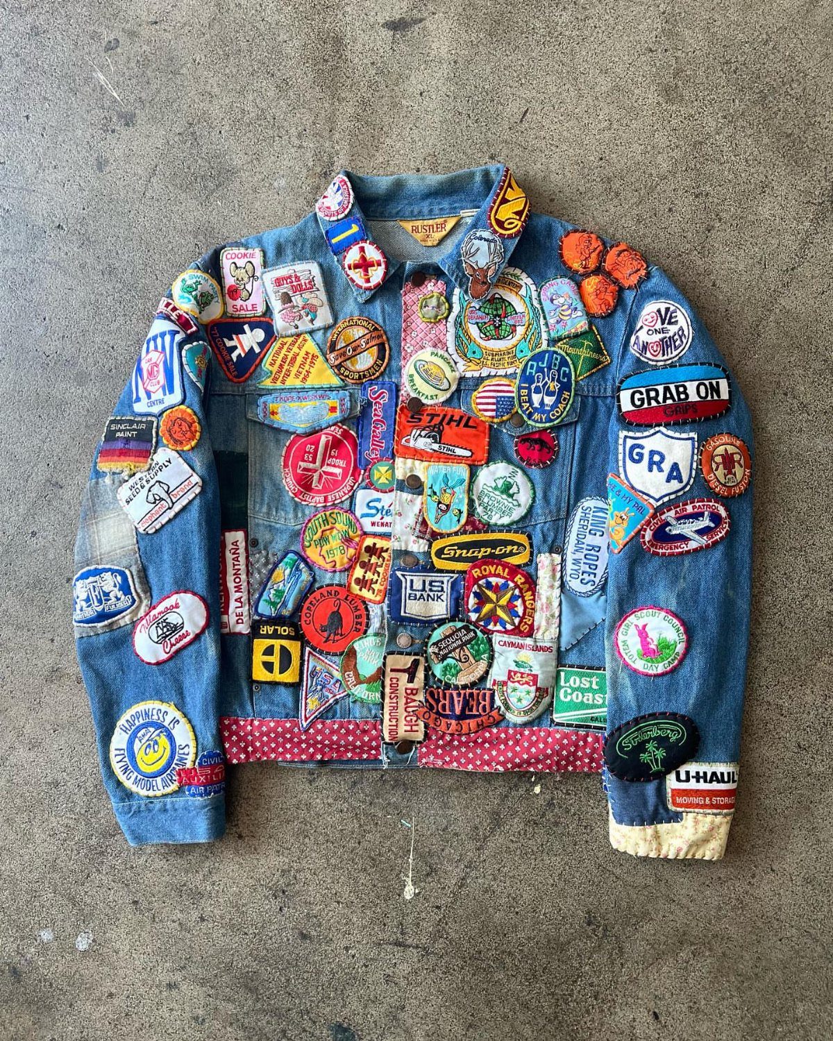 A blue denim jacket covered in many colorful vintage embroidered patches lying on concrete.