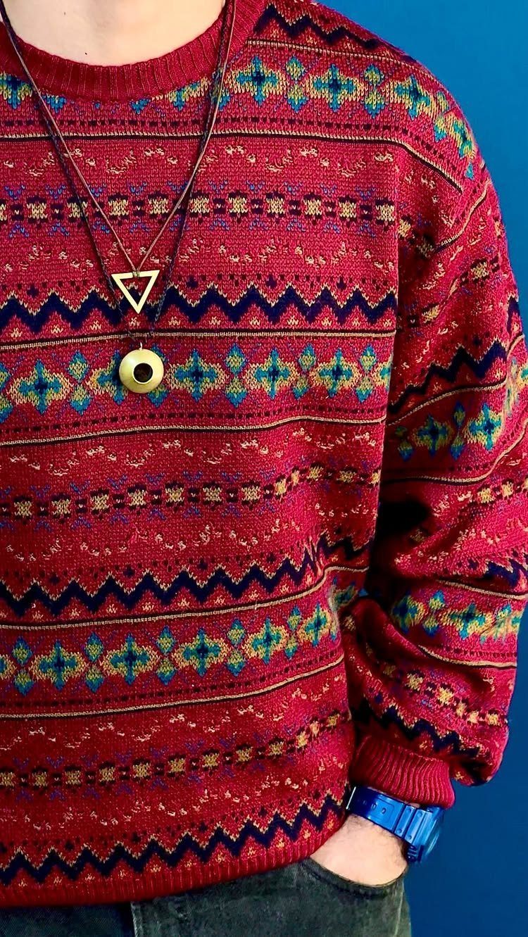 A person wearing a red patterned ribbed sweater and dark jeans against a blue background.