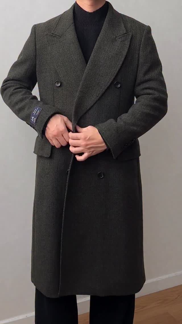 Man wearing a long, dark green herringbone patterned double-breasted coat over a black turtleneck.