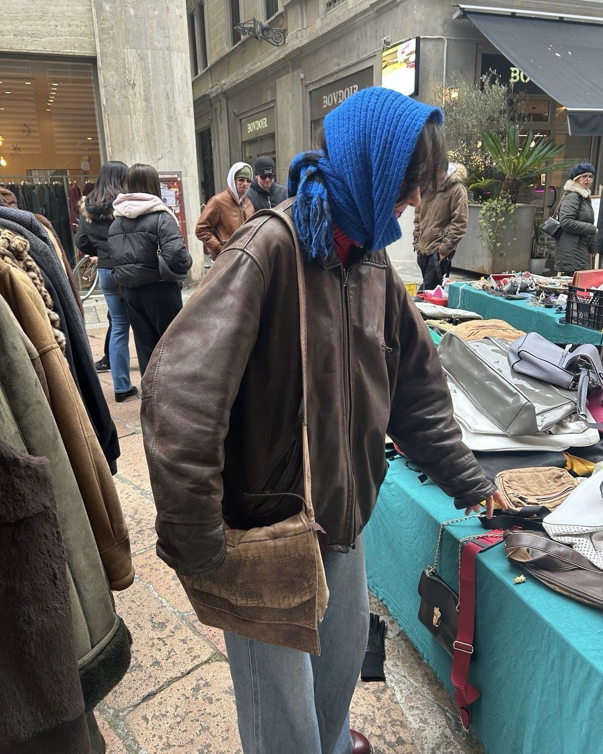 A person in a brown leather jacket and blue jeans browsing an outdoor market stall.