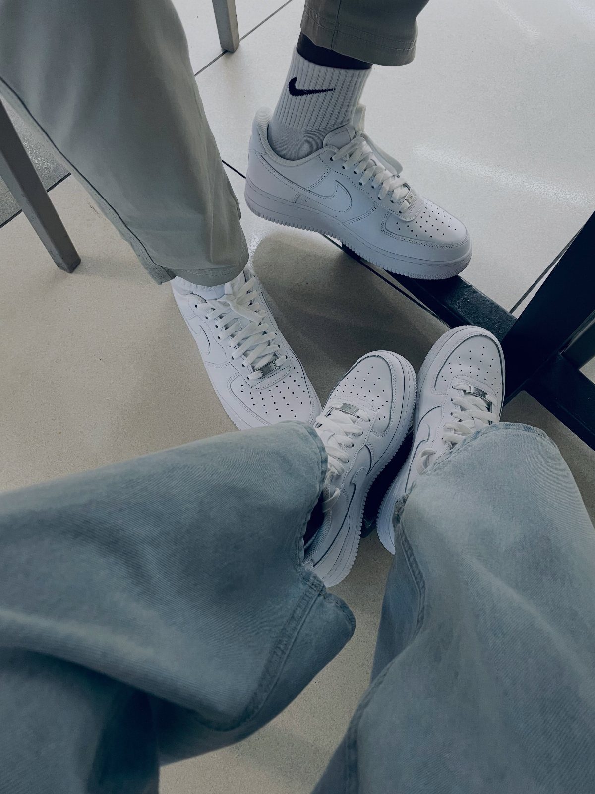 Three people wearing light blue denim jeans and white sneakers standing on a tiled floor.