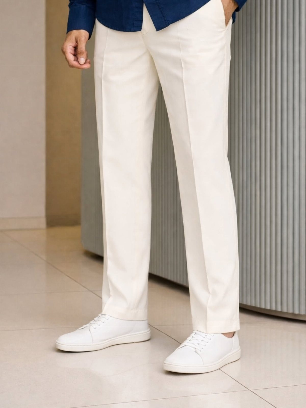 Person wearing cream tailored trousers and white leather sneakers standing on a tiled floor.