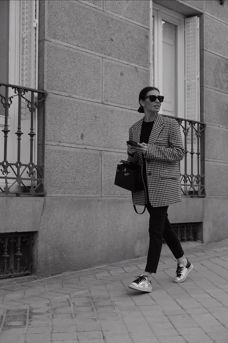 Woman walking in a patterned blazer, black pants, and white sneakers on a city sidewalk.
