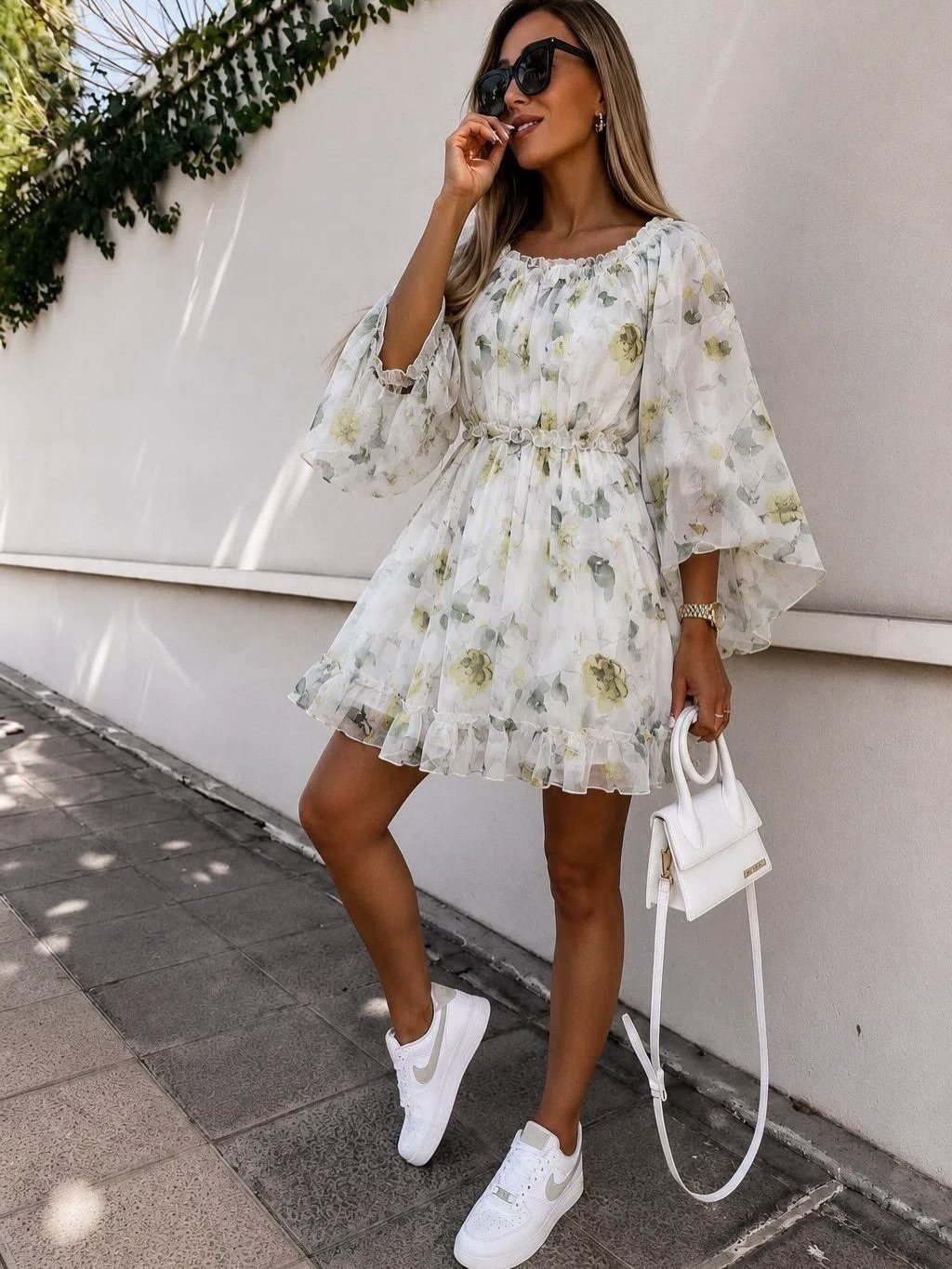 Woman in a white floral mini dress and white sneakers posing against a wall.