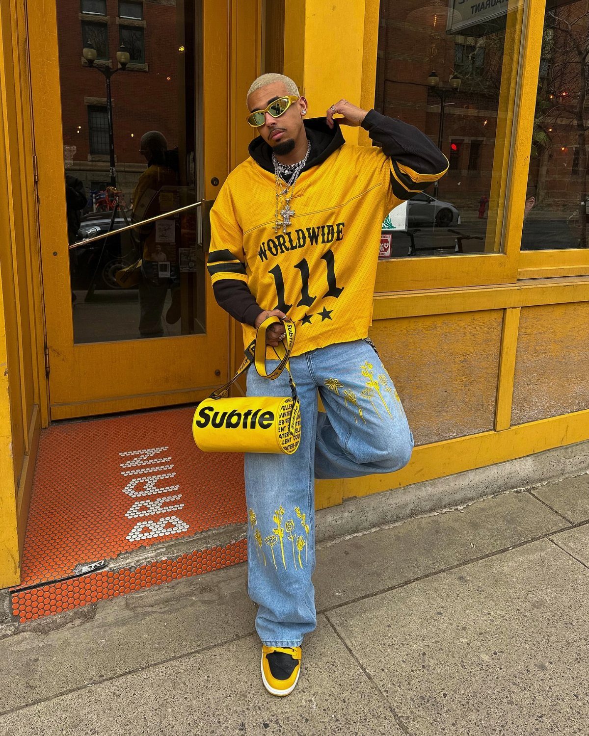 Man wearing a yellow and black graphic hoodie, blue jeans, and yellow accessories standing outside.