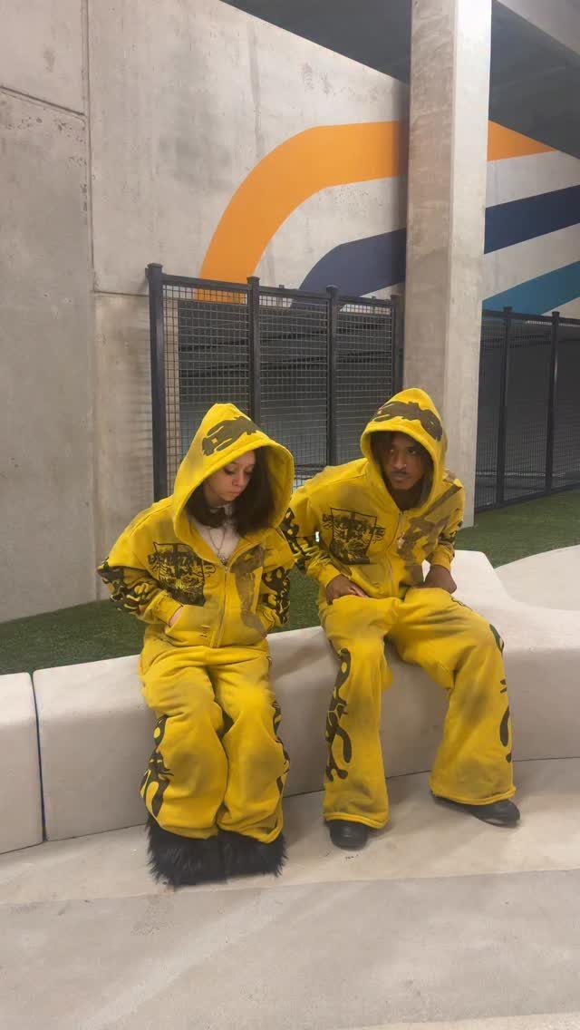Two people sitting outdoors wearing matching yellow graphic distressed hoodies and pants.