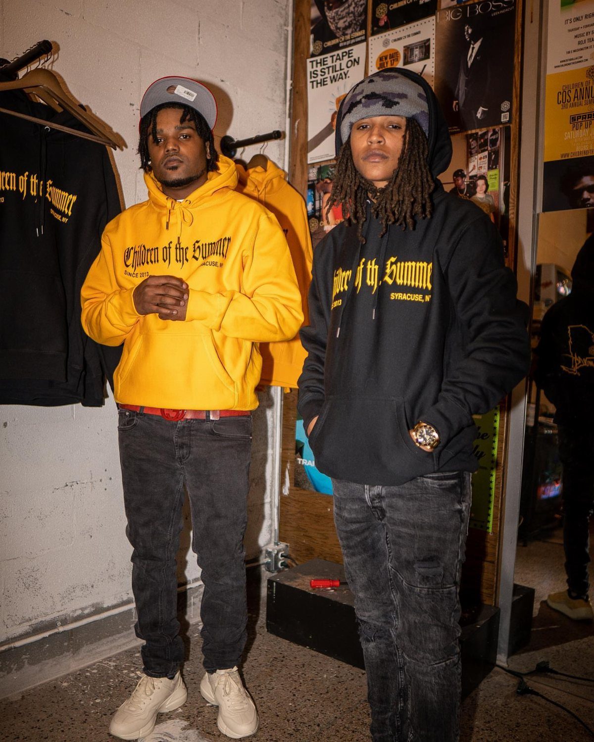 Two men posing while wearing yellow and black graphic hoodies and dark denim jeans.