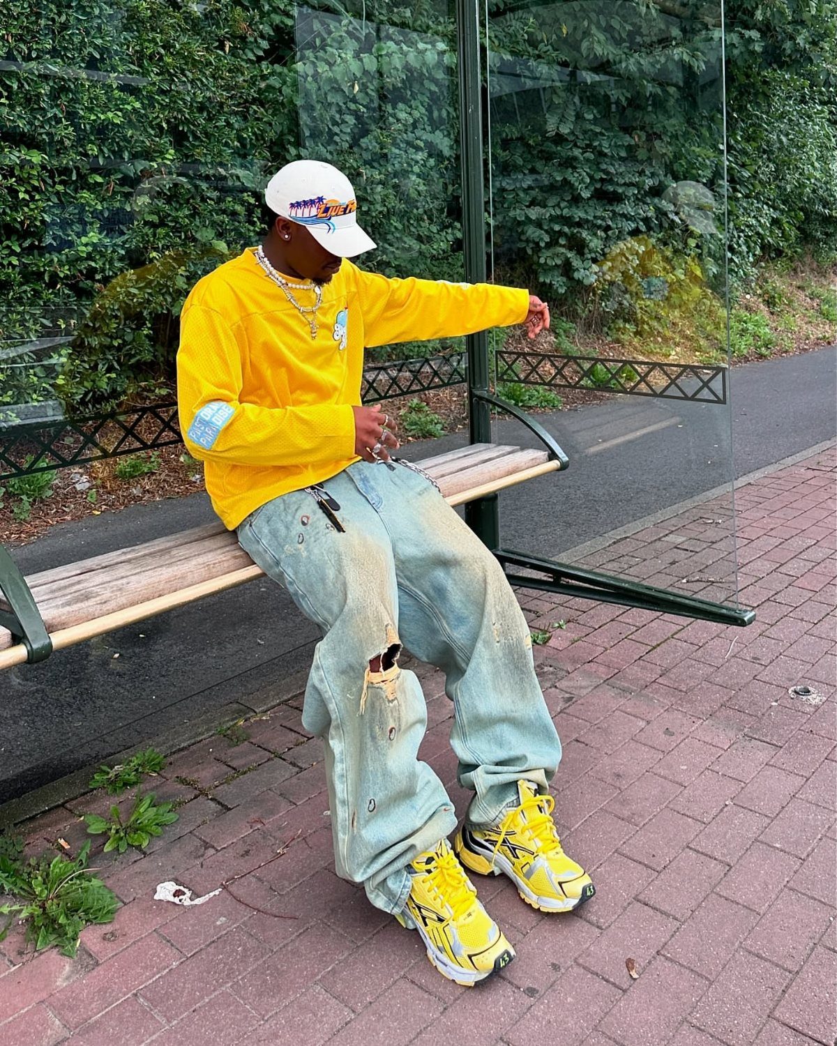 A person wearing a yellow long-sleeved shirt, light blue ripped jeans, and yellow sneakers sitting on a bench.