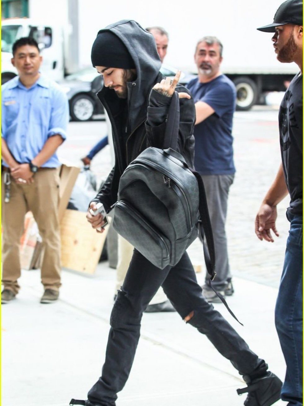 Man in black streetwear walking outdoors wearing a black hoodie, ripped jeans, and chunky sneakers.