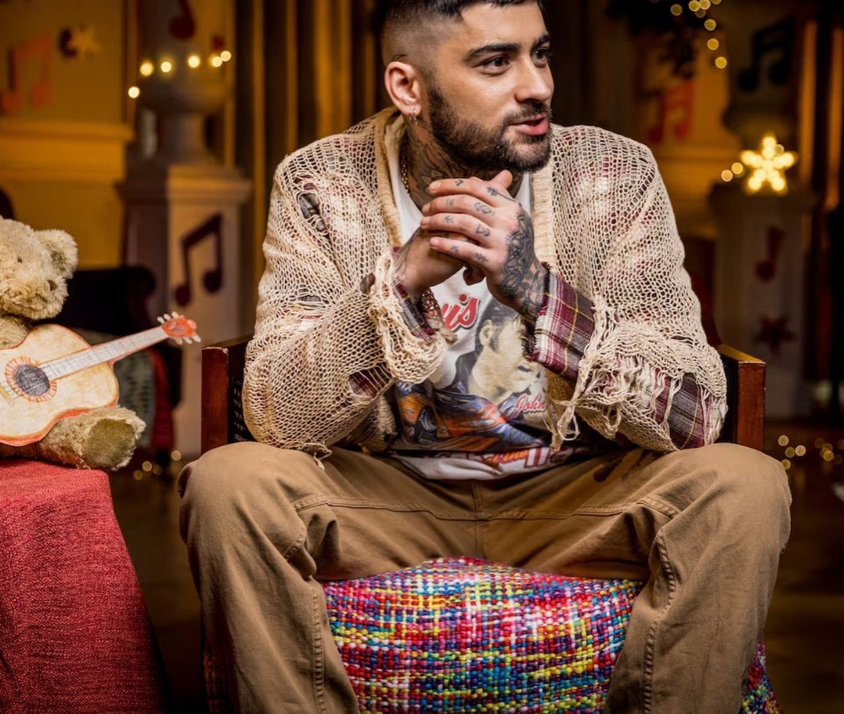 Man with tattoos sitting in a chair wearing a distressed knit cardigan and graphic t-shirt.
