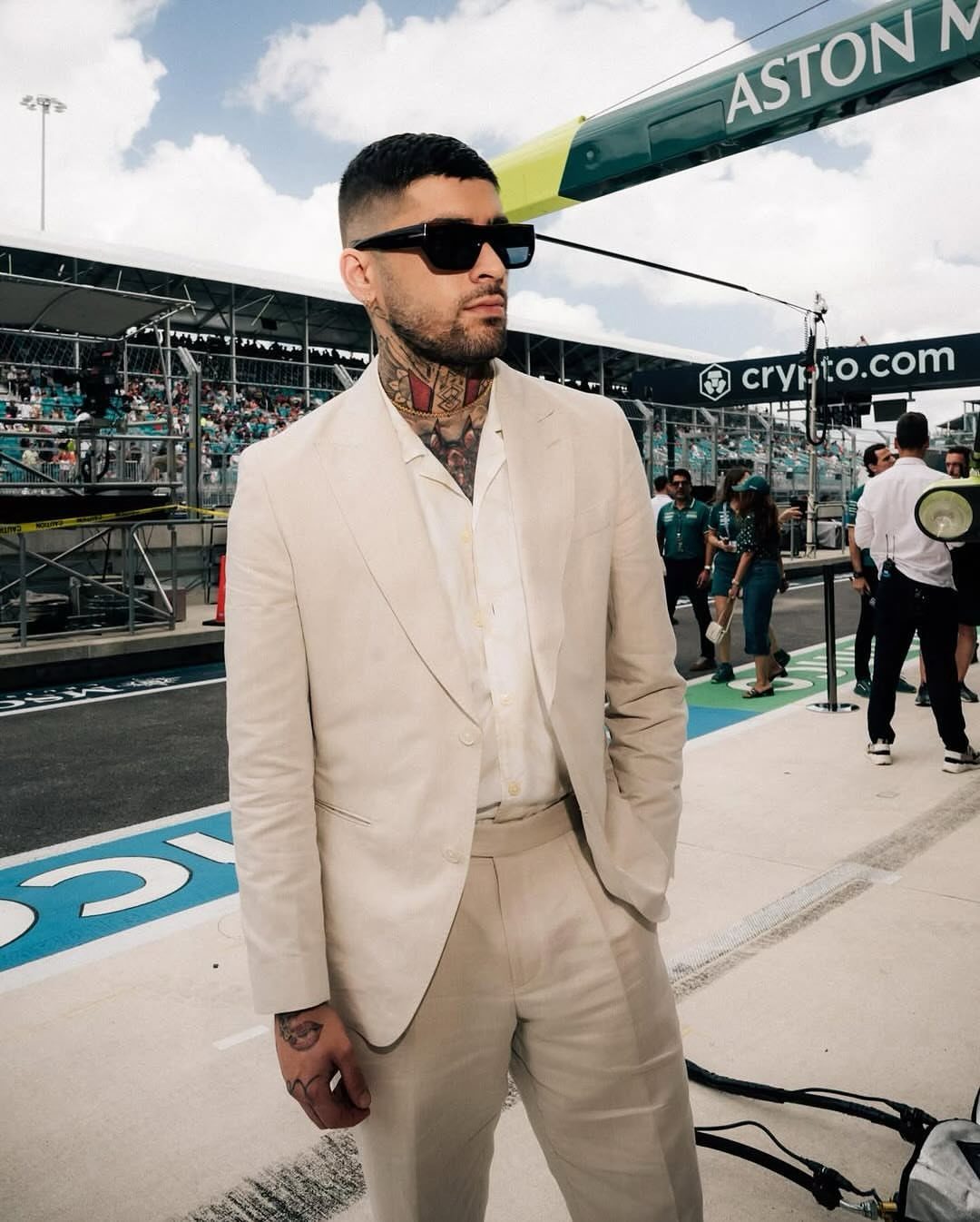 Man with neck tattoos wearing a beige suit and sunglasses at a racetrack.