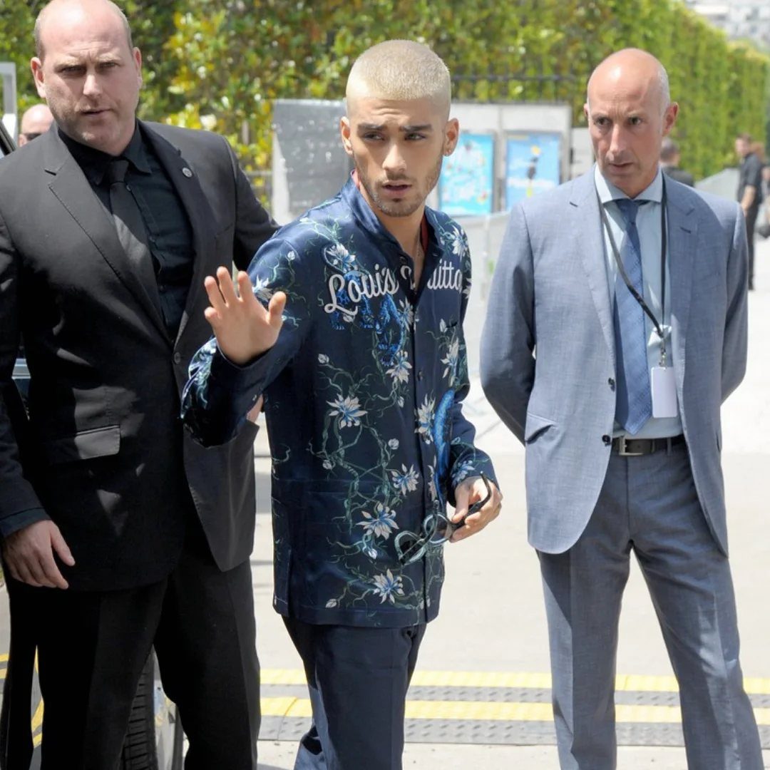 Zayn Malik walking outdoors wearing a dark blue Louis Vuitton floral patterned travel jacket.