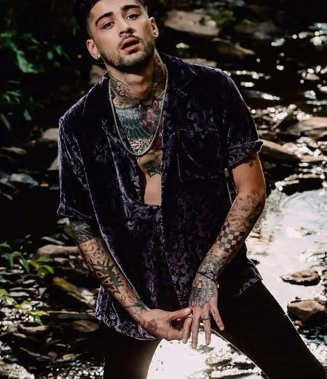 Tattooed man in a dark purple velvet shirt posing by a rocky stream.