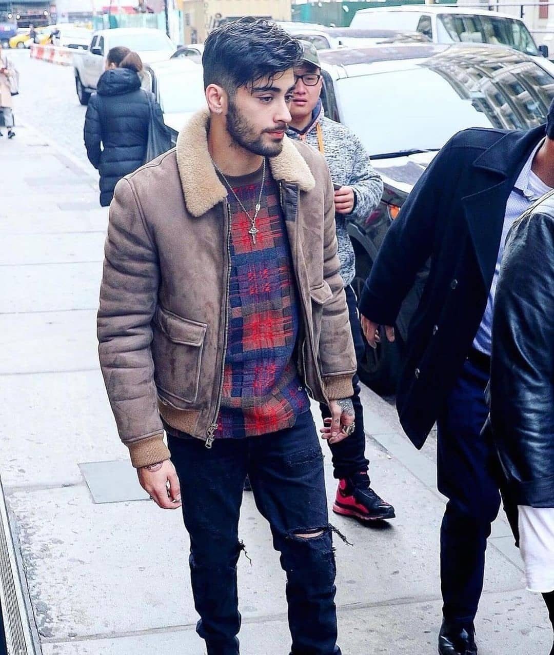 Man in a brown shearling jacket and plaid shirt walking on a city sidewalk.