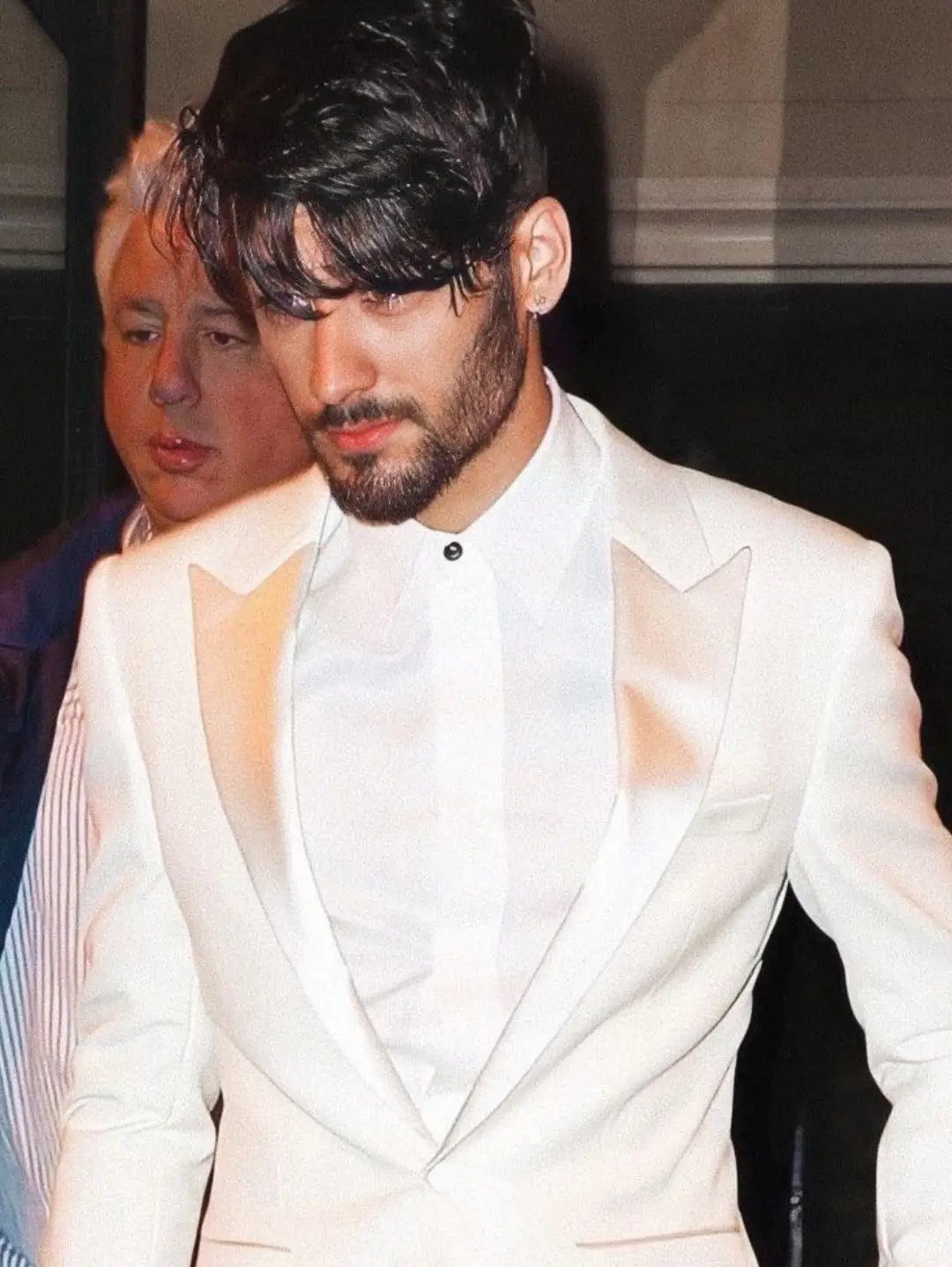 Man with dark hair and beard wearing a white suit jacket and shirt.